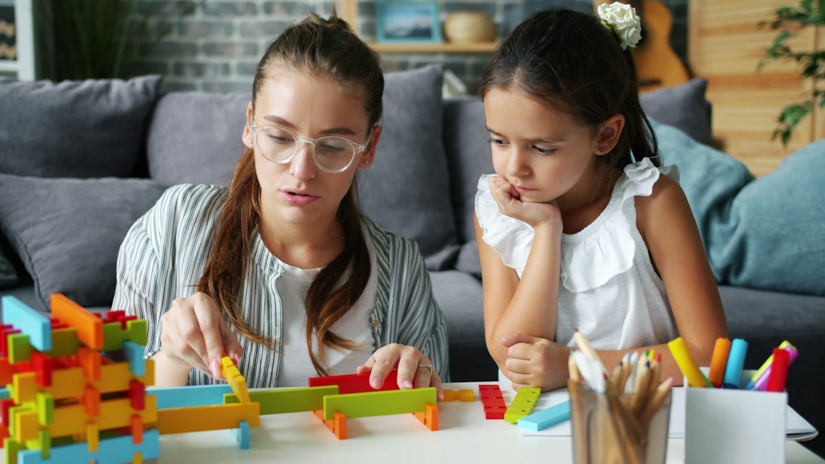 Cute little girl playing at home with caring mother having fun with wooden toys enjoying family activity. Childhood, motherhood and apartment concept.